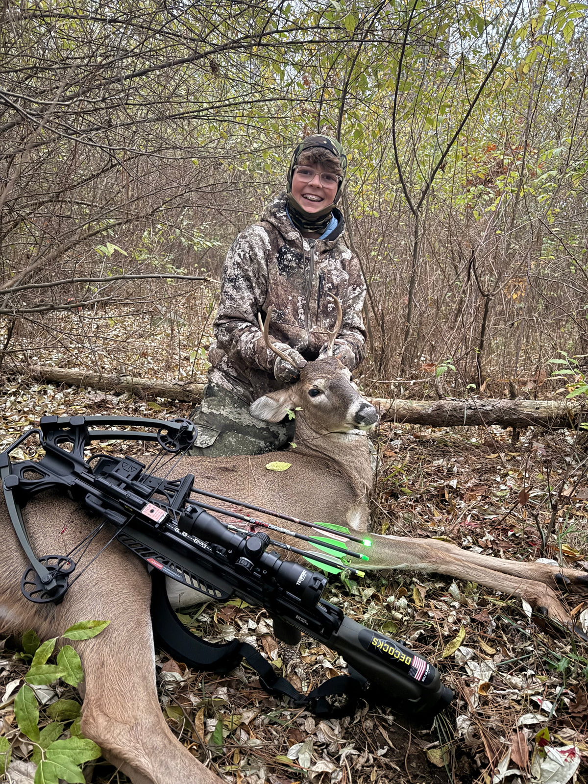 A crossbow hunter kneeling with a harvested whitetail in the hardwoods